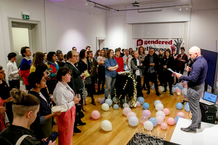 A crowd of varied queers stands around watching a man in a blue sweatshirt read off some notes. There are wine glasses and balloons in the forground, and a big Gendered Intelligence banner