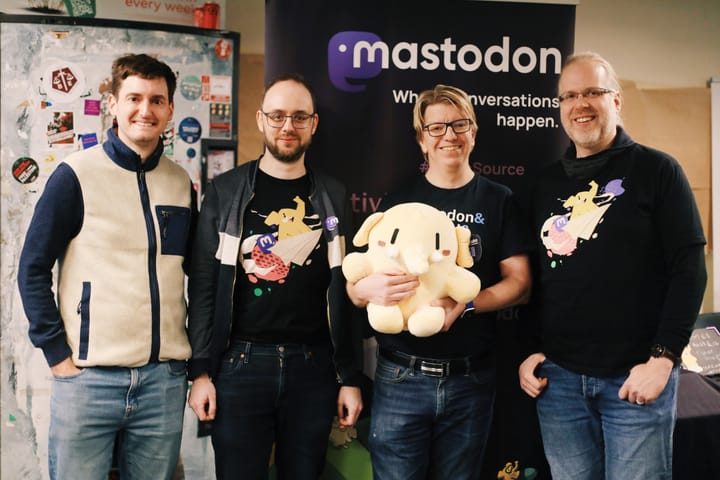 Four smiling white men face the camera wearing jackets, jeans and t-shirts with the Mastodon logo. Andy, interviewed in this piece, is holding a cute walrus plushie
